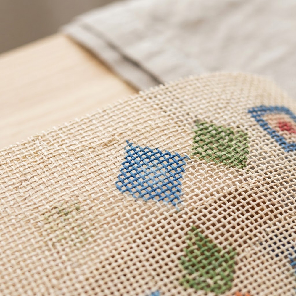 A close-up view of a woven needlepoint canvas showing the blank mesh and a few small, partially stitched diamond shapes in blue and green.